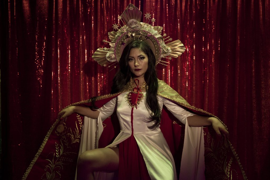 Actriz Magaly Solier posando como Lina de Lima con el fondo de tela roja y un vestido blanco con maquillaje y accesorios llamativos