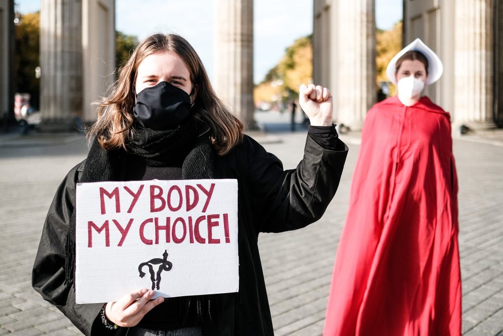 Una mujer vestida de negro sostiene un carte que dice "my body, my choice" y otra parada detrás de ella está vestida como en el Cuento de la Criada